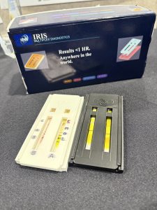 A prototype of diagnostic testing cassettes, one black and one white, in front of a navy box with the IRIS logo.